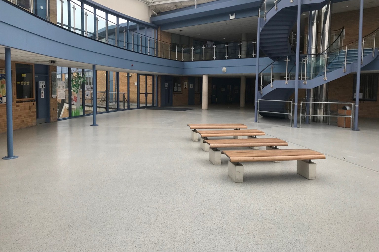 School atrium with decorative resin flooring, wooden benches, blue railings, and a spiral staircase.