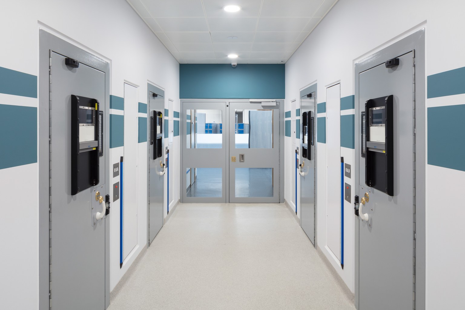 Light resin flooring through a police custody corridor with secure cell doors.