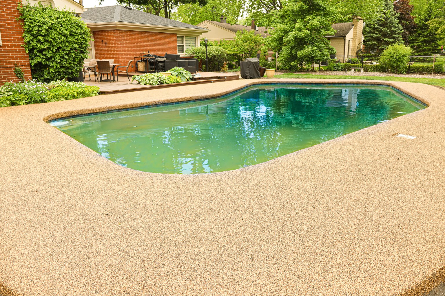 Tan rubber-aggregate pool deck surrounding a backyard swimming pool beside a brick home.