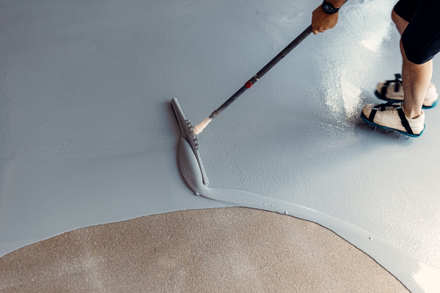 Installer in spiked overshoes spreading gray epoxy onto concrete with a long-handled squeegee.