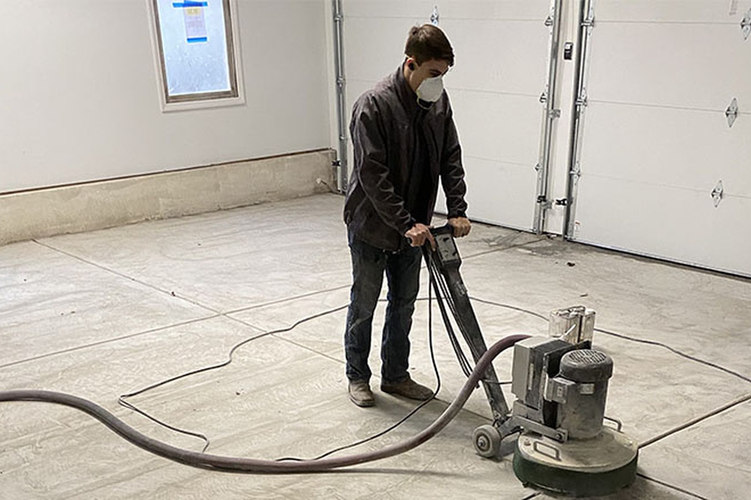 Technician diamond-grinding a bare concrete garage floor with dust extraction.
