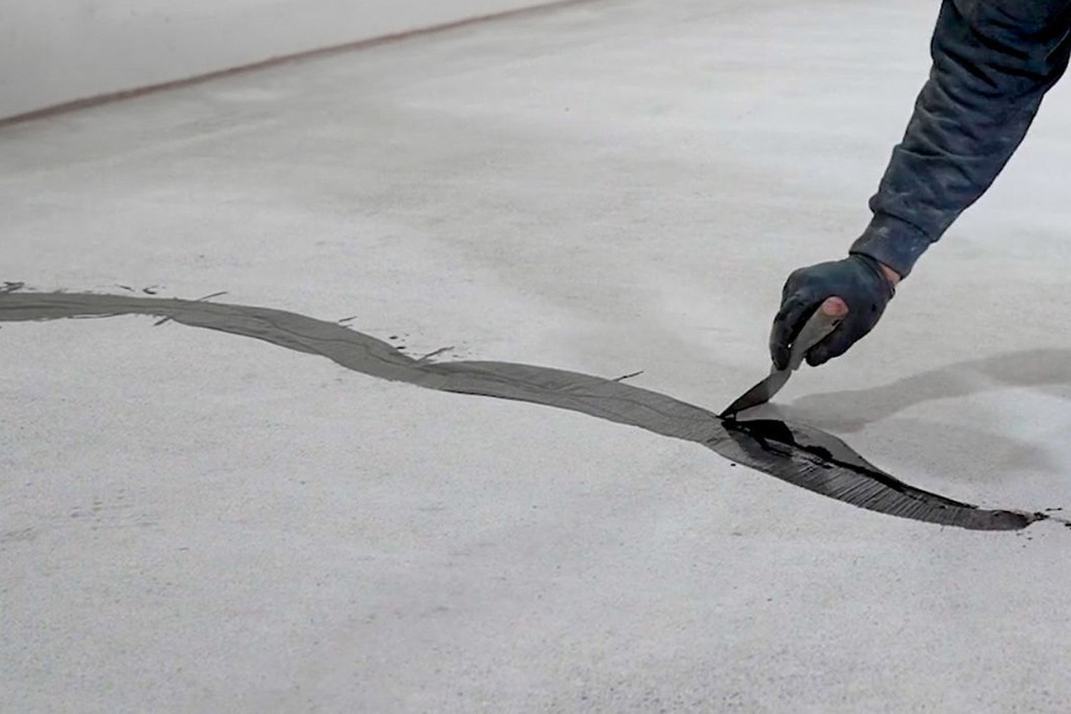 Worker smoothing dark repair filler into an irregular concrete-floor crack with a putty knife.